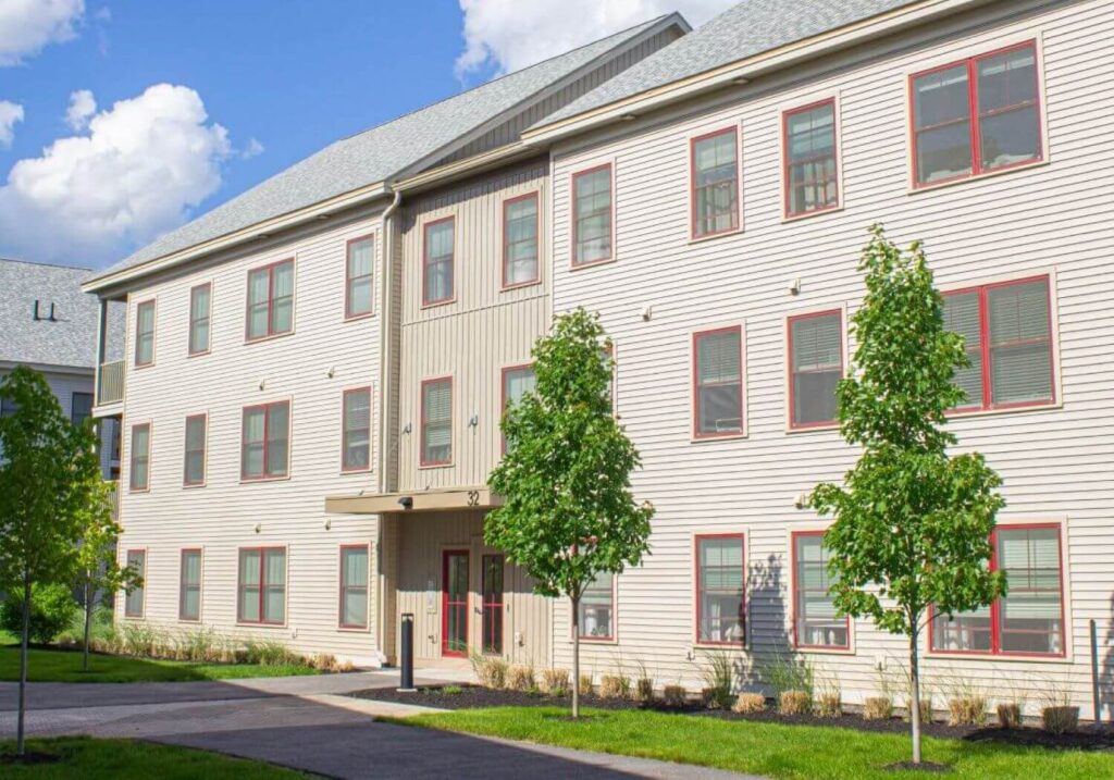 Two Frontrunner Condos buildings at The Downs in Scarborough, Maine, featuring brown siding, red window trim, landscaped trees, and a clear blue sky.