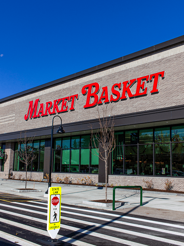 Exterior view of the new Market Basket grocery store at The Downs in Scarborough, Maine, scheduled to open in November 2026. The storefront features a large red Market Basket sign, expansive glass windows, and a pedestrian walkway beneath a clear blue sky.